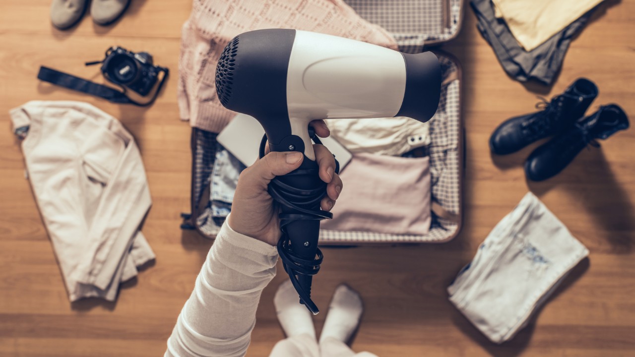 Person holding a hair dryer with their open suitcase filled with clothes on the ground below.