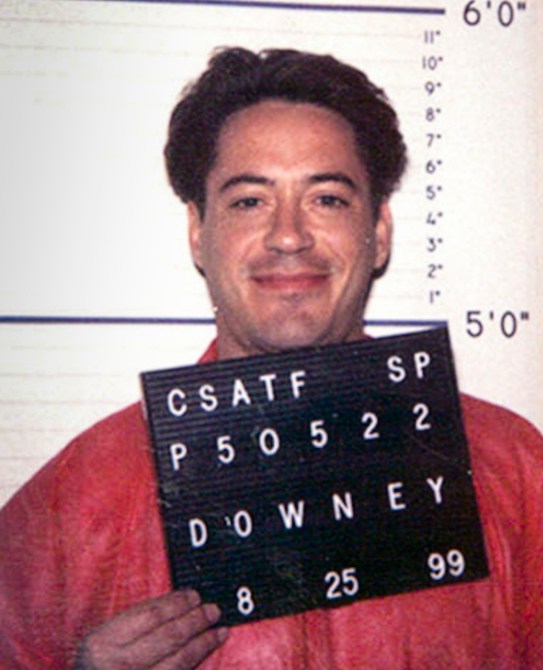 In a orange shirt, Robert Downey Jr smiles fro his mugshot.