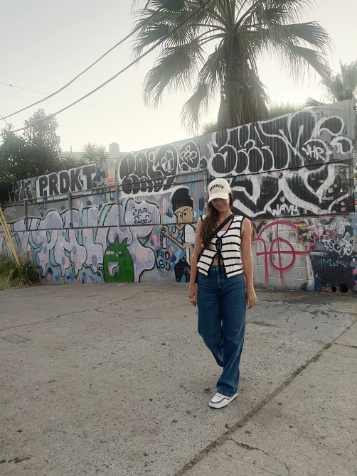 A woman in a white baseball hat, striped tank, and blue jeans in front of a graffiti wall
