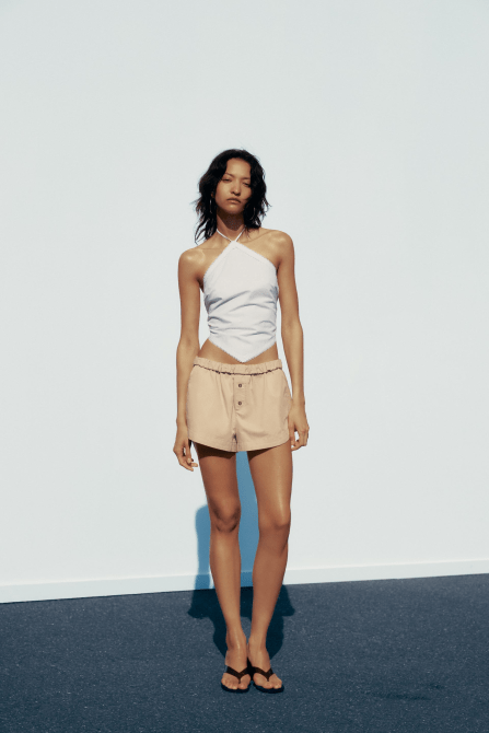A model wearing the Zara Fold-Over Waist Mini Shorts in beige with a white halterneck top.